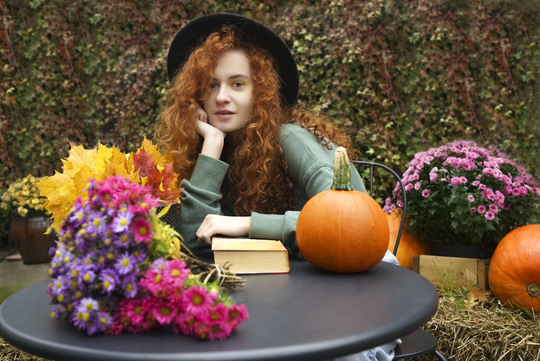 dreaming young woman reading a romantic book in a cozy warm atmosphere. redhead Lady enjoying the autumn mood. black table with yellow maple flowers and orange pumpkins.  - 写真・画像