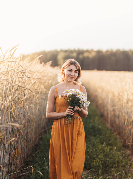 A young beautiful girl stands on a wheat field and holds a bouquet of daisies in her hands. - Photo, image