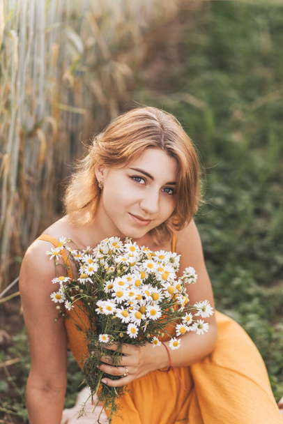 Una giovane bella ragazza siede su un campo di grano e tiene in mano un mazzo di margherite. - Foto, immagini