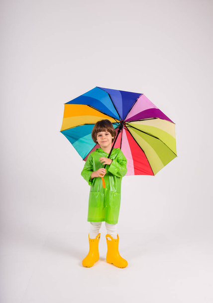 een kleine jongen in een groene regenjas en gele rubberen laarzen staat en houdt een veelkleurige paraplu op een witte achtergrond met een plaats voor tekst - Foto, afbeelding