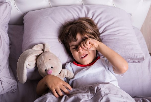 Ένα μικρό παιδί με πιτζάμες ξυπνάει σε ένα κρεβάτι με λούτρινο παιχνίδι. - Φωτογραφία, εικόνα