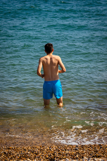 A 18 year old Caucasian shirtless teenage boy in swimwear walking into the sea - Foto, imagen