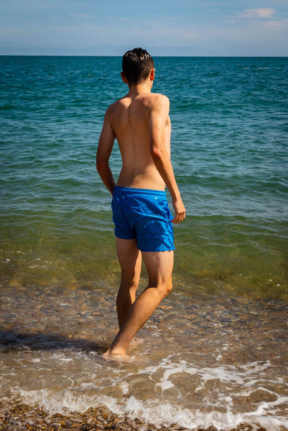 A 18 year old Caucasian shirtless teenage boy in swimwear walking into the sea - Fotografie, Obrázek