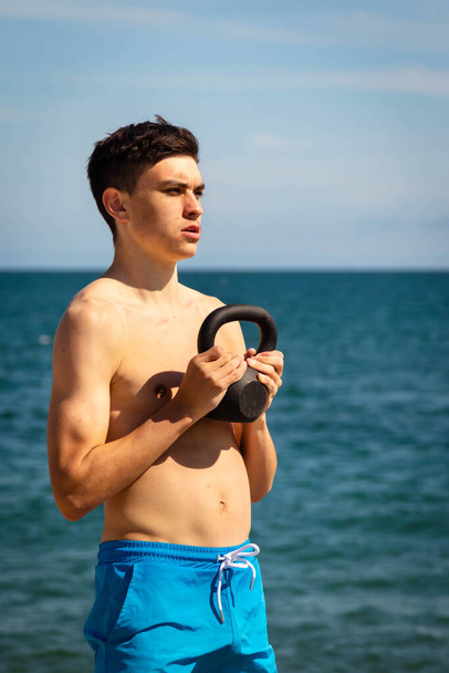 A 18 year old Caucasian shirtless teenage boy on a beach exercising with a kettlebell weight - Фото, изображение