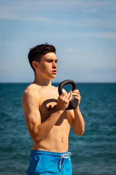 A 18 year old Caucasian shirtless teenage boy on a beach exercising with a kettlebell weight - Photo, image