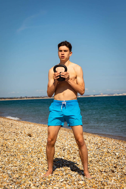 A 18 year old Caucasian shirtless teenage boy on a beach exercising with a kettlebell weight - Fotografie, Obrázek