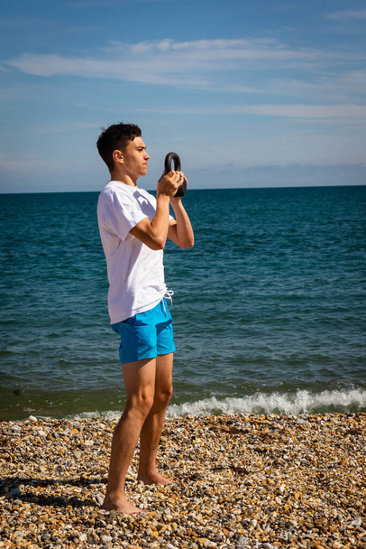 A 18 year old Caucasian shirtless teenage boy on a beach exercising with a kettlebell weight using both hands - Photo, image