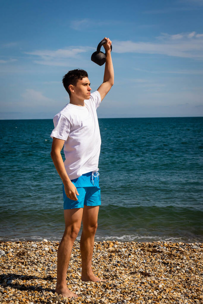 A 18 year old Caucasian teenage boy on a beach exercising with a kettlebell weight - Foto, imagen