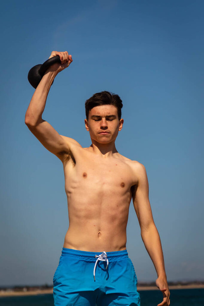 A 18 year old Caucasian shirtless teenage boy in swimwear exercising with a kettlebell weight - 写真・画像
