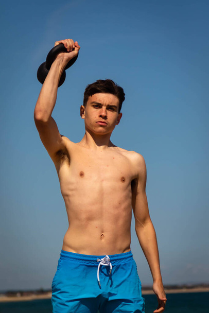 A 18 year old Caucasian shirtless teenage boy in swimwear exercising with a kettlebell weight - Foto, Bild