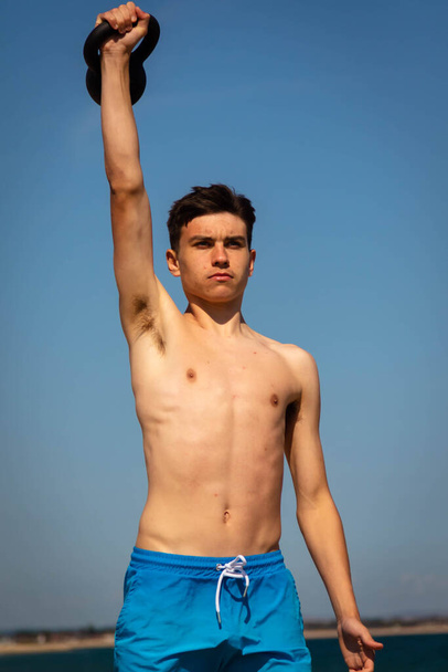 A 18 year old Caucasian shirtless teenage boy in swimwear exercising with a kettlebell weight - Фото, зображення