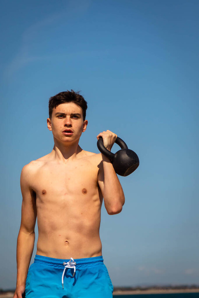 A 18 year old Caucasian shirtless teenage boy in swimwear exercising with a kettlebell weight - 写真・画像
