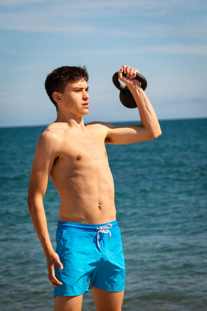 A 18 year old Caucasian shirtless teenage boy in swimwear exercising with a kettlebell weight - Foto, afbeelding