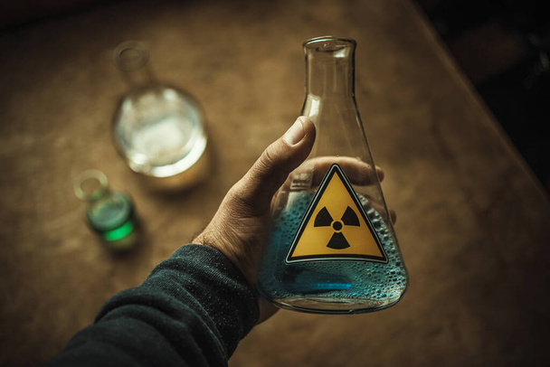 Рука держит стеклянную фляжку в лаборатории с голубой жидкостью. Желтая наклейка на стекле предупреждает о радиации. Лабораторные испытания. - Фото, изображение