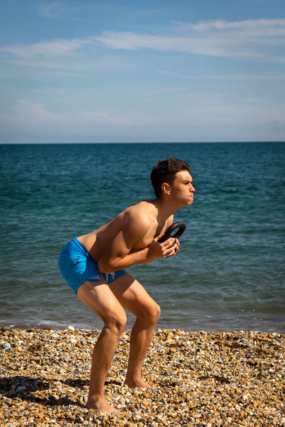 A 18 year old Caucasian shirtless teenage boy on a beach exercising with a kettlebell weight - Foto, imagen