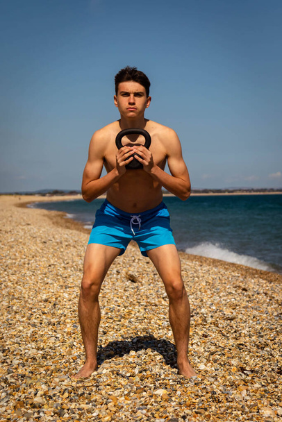 A 18 year old Caucasian shirtless teenage boy on a beach exercising with a kettlebell weight - Foto, afbeelding