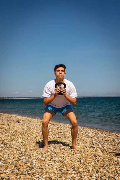 A 18 year old Caucasian teenage boy on a beach exercising with a kettlebell weight - Fotó, kép