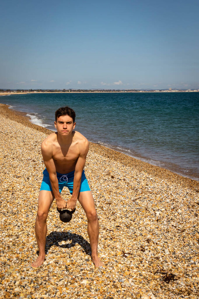 A 18 year old Caucasian shirtless teenage boy on a beach exercising with a kettlebell weight - Φωτογραφία, εικόνα