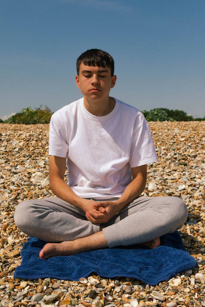 A Caucasian teenage boy meditating on a stony beach with his hands resting on his lap - Фото, изображение