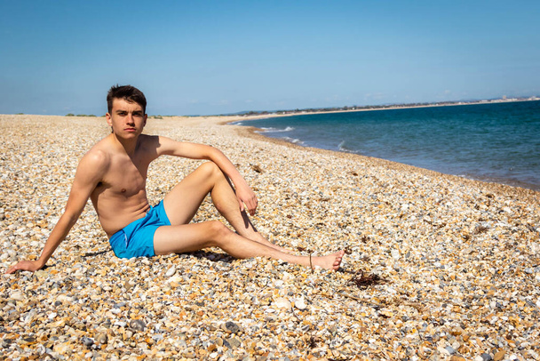 A 18 year old Caucasian shirtless teenage boy relaxing on a beach next to the sea - Zdjęcie, obraz