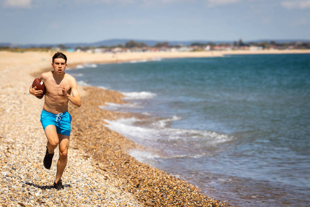 A 18 year old Caucasian shirtless teenage boy running on a beach with an American Footbal - Foto, afbeelding