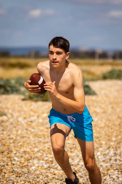 A 18 year old Caucasian shirtless teenage boy running on a beach with an American Football - Zdjęcie, obraz