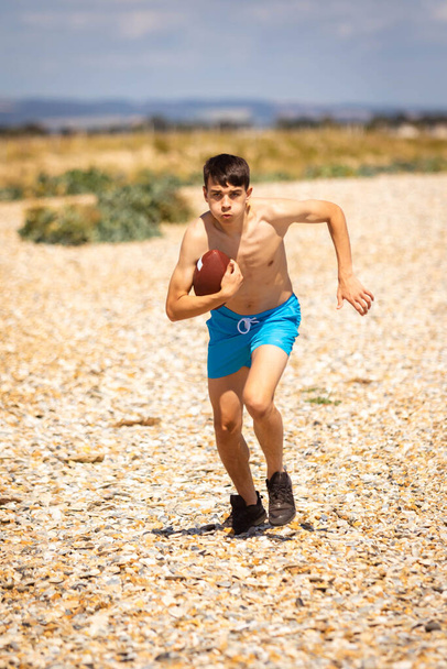 A 18 year old Caucasian shirtless teenage boy running on a beach with an American Football - Foto, Bild