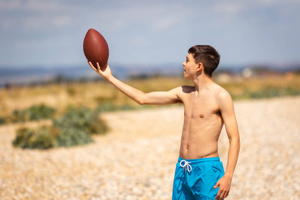 A 18 year old Caucasian shirtless teenage boy playing on a beach with an American Football - Photo, image