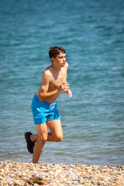 A 18 year old Caucasian shirtless teenage boy running on a beach next to the sea - Фото, изображение