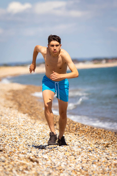 A 18 year old Caucasian shirtless teenage boy running on a beach next to the sea - 写真・画像
