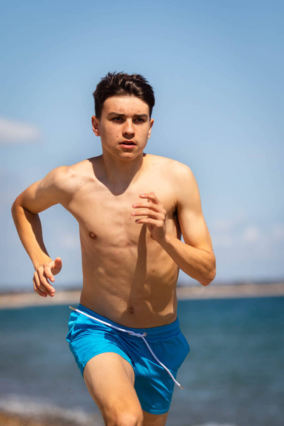 A 18 year old Caucasian shirtless teenage boy running on a beach next to the sea - Zdjęcie, obraz