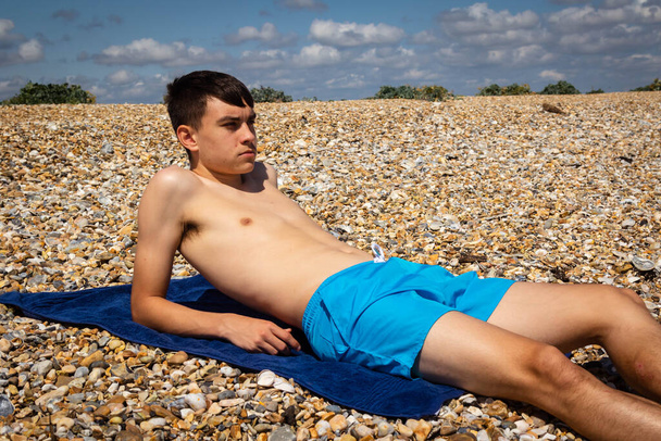 A 18 year old Caucasian shirtless teenage boy sunbathing on a stoney beach - Фото, изображение
