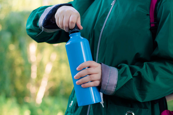 A girl in a green jacket is holding in her hands a shining hiking bottle for water. Pink backpack. Hike in the woods. - Foto, immagini