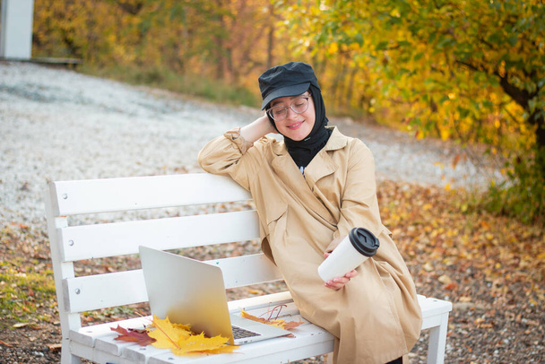 A Muslim woman in a hijab in a beige trench coat sits on a bench with a cup of coffee and a laptop in the park. Muslim business woman. Student. Glasses for sight. - Foto, immagini