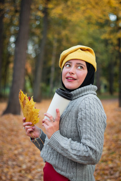 Young Muslim woman in a yellow hat with a gray sweater with a cup of coffee and a yellow autumn leaf. Autumn Park. Femininity and a smile - Foto, Imagem