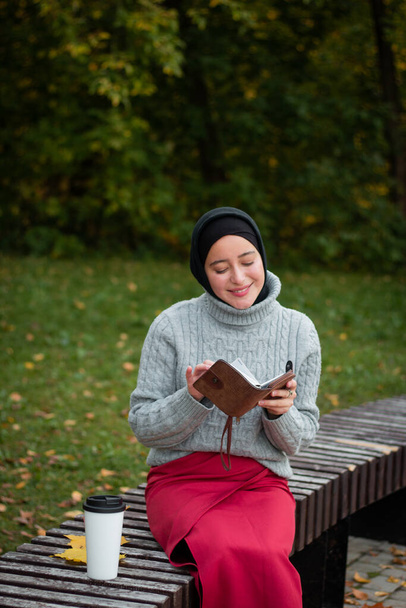 A young Muslim woman in a gray sweater sits with a notebook on a bench. Cup of coffee and autumn leaves. Student. Education. University student - Foto, afbeelding