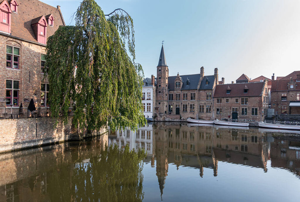 Brujas, Flandes, Bélgica - 4 de agosto de 2021: El tranquilo canal Dijver refleja la piedra marrón Huidevettershuis y los barcos bajo el cielo azul claro y el follaje verde a un lado. - Foto, Imagen