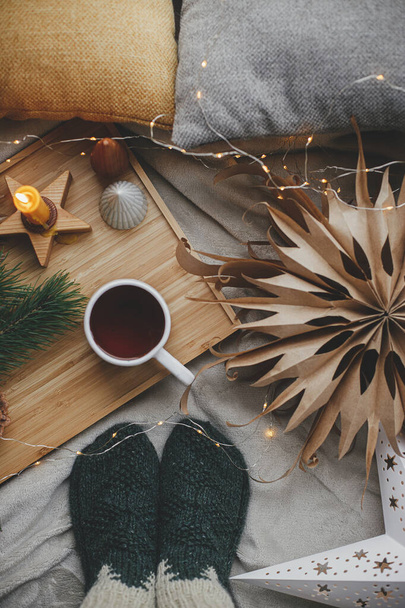 Pieds de femme en chaussettes de laine sur lit doux avec tasse de thé chaude, étoiles de Noël, lumières dorées, arbres, bougie et oreillers. Vue de dessus. Moments confortables à la maison scandinave. Vacances d'hiver et d'automne - Photo, image