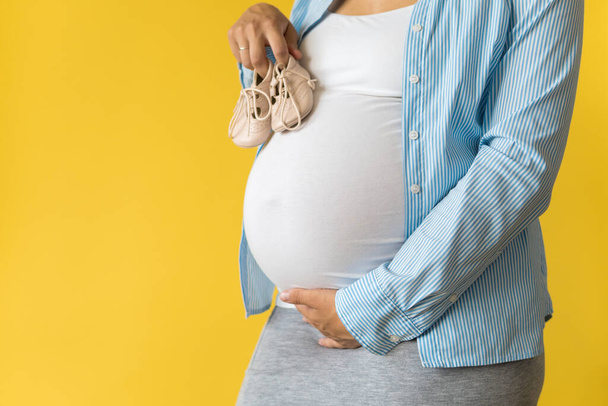 Motherhood, femininity, love, care, waiting, hot summer - bright croped Close-up unrecognizable pregnant woman in shirt with small baby shoes hand over tummy rub belly on yellow background, copy space - 写真・画像