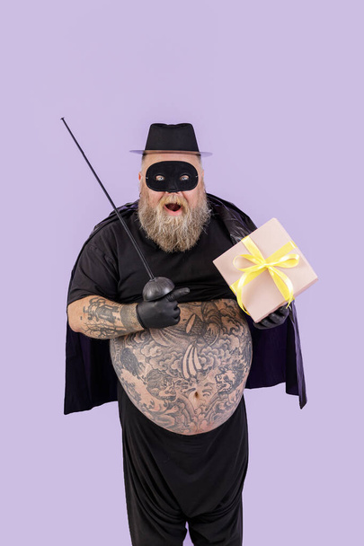 Surprised mature man with overweight in Zorro suit points onto gift box on purple background - Foto, Bild