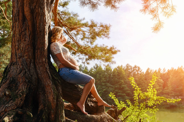 Młoda ciężarna kobieta siedzi na korzeniach pod drzewem nad rzeką - Zdjęcie, obraz