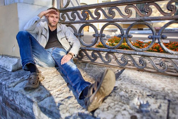 European man in jeans sits on road outdoors. - Фото, изображение