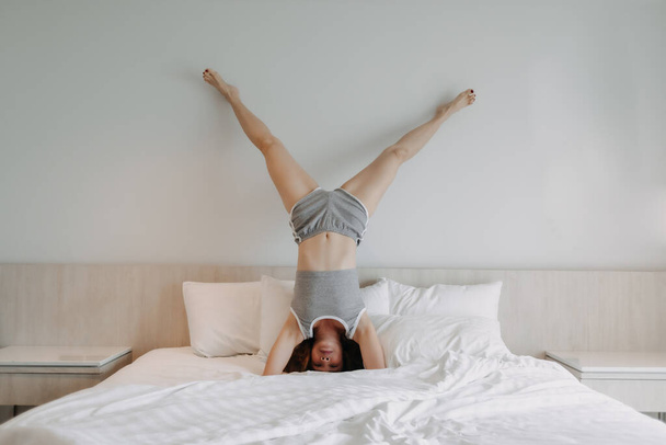 Playful woman is doing upside down pose on the bed. - Φωτογραφία, εικόνα