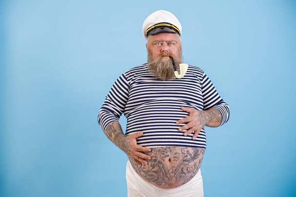 Middle aged obese man in sailor costume holds hands on tummy on light blue background - Photo, Image