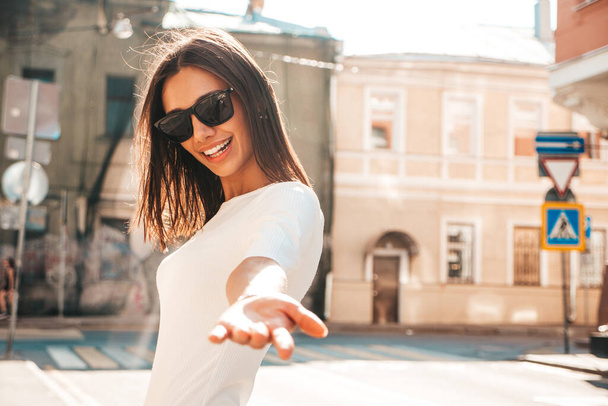 Young beautiful smiling hipster woman in trendy summer sundress. Sexy carefree woman posing on the street background at sunset. Positive model in sunglasses. Outstretching hand, asking to follow her - Photo, Image