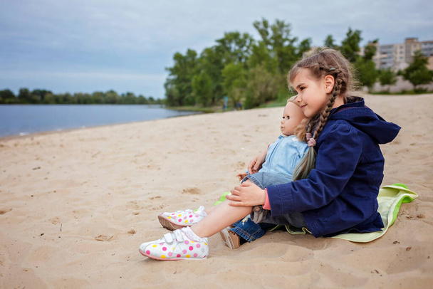 Самотня чарівна вісім років дівчина грає з лялькою на осінньому пляжі, мати і дочка гра
 - Фото, зображення