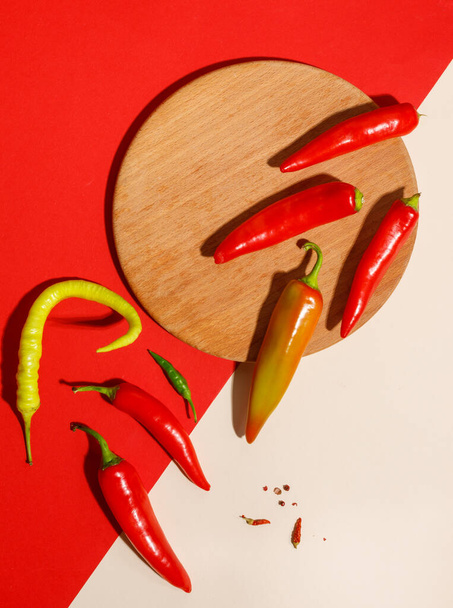 Horké červené a zelené papriky v lusky na červeném a béžovém pozadí a kulaté dřevěné desky. Horní pohled, svislý - Fotografie, Obrázek