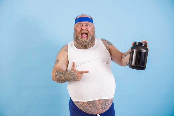 Joyful obese man presenteert grote fles voedingsstof supplement op lichtblauwe achtergrond - Foto, afbeelding