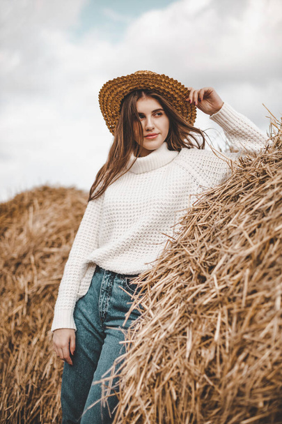 Een mooi plattelandsmeisje in een witte trui, jeans en een strohoed staat naast een hooiberg. - Foto, afbeelding