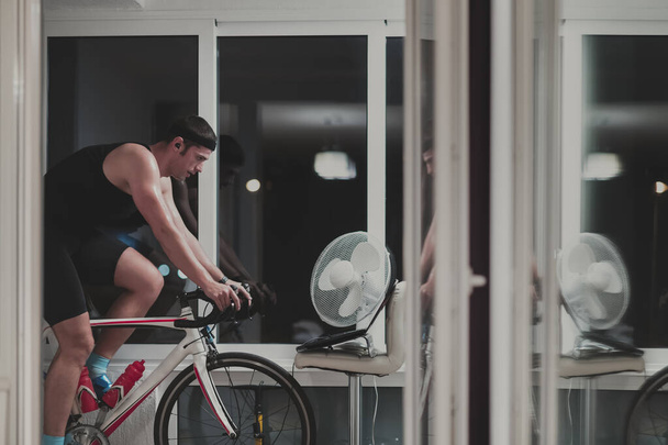 Man cycling on the machine trainer he is exercising in the home at night playing online bike racing game - Zdjęcie, obraz
